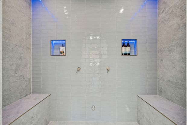 A contemporary tiled bathroom shower or wet-room enclosure, viewed straight on. The walls are clad in glossy pale grey-white rectangular tiles, with two recessed illuminated niches containing toiletries. Brushed metal shower controls are mounted centrally, and built-in tiled benches frame the lower left and right sides. Blue LED lighting adds a cool accent.