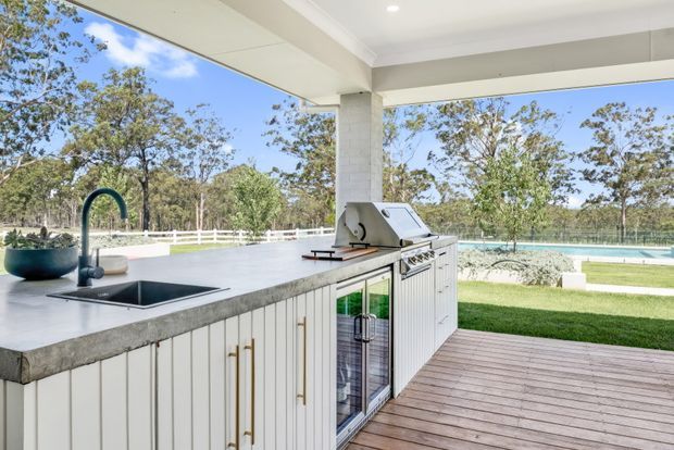 A covered outdoor entertaining area with a built-in outdoor kitchen overlooking a landscaped lawn and swimming pool. The kitchen features a large concrete-look island with integrated sink, white vertical-panel cabinetry with brass-toned handles, a glass-front beverage refrigerator, and a stainless-steel barbecue. Timber decking extends around the space, with expansive open glazing framing the garden and pool.
