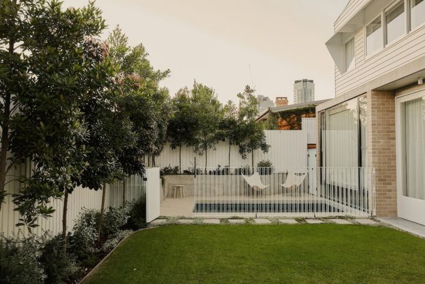 A compact landscaped courtyard beside a contemporary multi-storey residence. The space features a neatly clipped lawn, mature screening trees and shrubs, a white slatted privacy fence, slim vertical metal balustrades, and a small rectangular plunge pool or water feature with suspended hammock-style seating. The house has pale horizontal cladding, light brickwork, large glazed doors and windows, and a clean modern composition. A distant high-rise is visible beyond the garden.