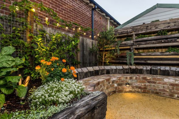 A compact landscaped courtyard garden with curved raised brick-and-timber seating, a textured rendered or gravel-like floor, dense planting, climbing greenery, warm integrated lighting, and a rustic timber privacy screen. A red-brick building wall and a small roof structure frame the space.