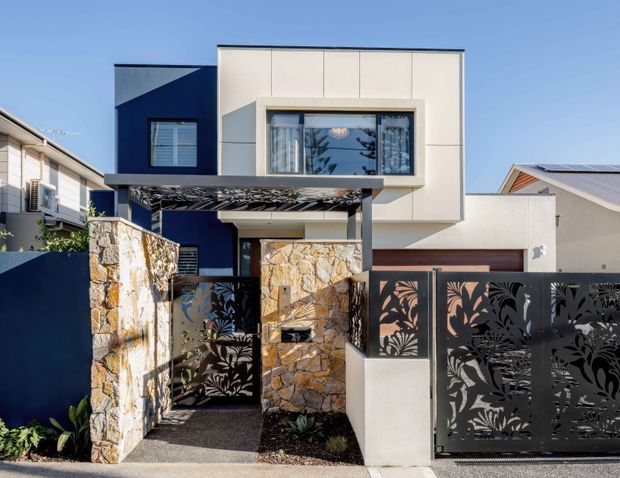 Street-facing view of a contemporary multi-storey residence with a crisp white upper facade, dark navy feature wall, projecting window box, flat roofline, and a covered entry canopy. The front boundary combines irregular natural-stone walls with decorative laser-cut metal gates featuring botanical patterns.