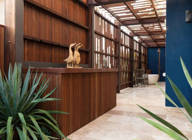 A refined covered outdoor courtyard or entry area with dark timber slatted screening, a substantial timber counter or built-in enclosure, pale stone-look paving, and a vivid blue wall. Tropical agave-like planting and a sculptural bird ornament add an organic decorative character. A louvred timber roof allows filtered daylight into the space, creating a warm, resort-like atmosphere.
