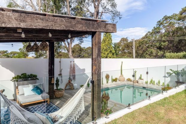 Bright outdoor poolside entertaining area with a rustic timber pergola, hanging pendant lights, hammock, cushioned outdoor seating, patterned rug, and potted plants. A glass pool fence encloses a compact swimming pool beside a white rendered boundary wall, with mature trees and blue sky beyond.