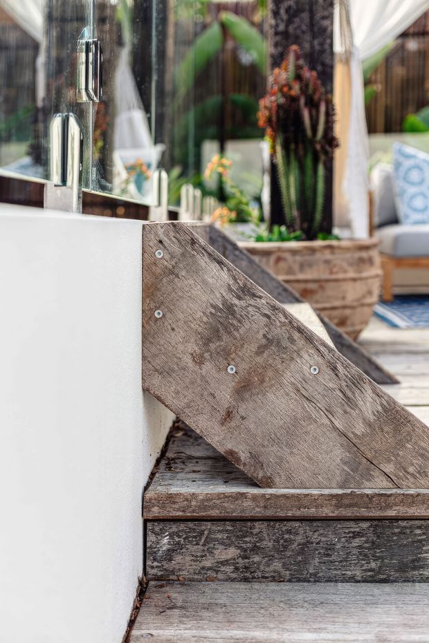 Close-up detail of a weathered timber outdoor staircase or raised deck edge beside a smooth white rendered wall. A triangular timber side support with exposed fasteners sits above aged timber treads and decking. Reflective glazing and a chrome outdoor wall fixture are visible to the left, while large planters, tropical planting, and a partially visible cushioned outdoor seating area appear in the background. The image has a bright, relaxed outdoor residential feel with rustic reclaimed timber detailing.