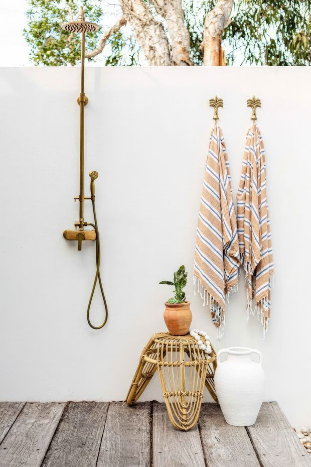 Bright outdoor shower vignette against a white rendered wall, featuring an antique brass shower fixture, two striped hanging towels, weathered timber decking, a woven rattan stool or basket, terracotta potted plant, and white ceramic vessel. The scene has a relaxed coastal, natural aesthetic with mature trees visible above the wall.