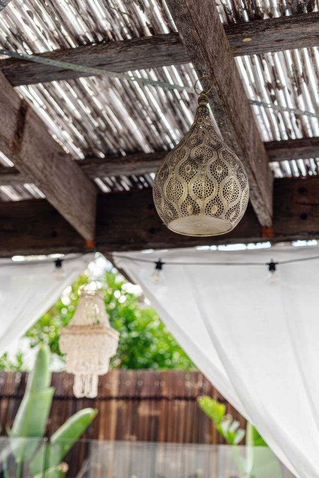 Close-up view of a rustic covered outdoor area with a weathered timber beam-and-rafter roof, corrugated translucent roofing panels, and a hanging ornate perforated metal lantern. White fabric curtains, string lights, tropical greenery, and a timber privacy fence create a relaxed, bohemian outdoor atmosphere.