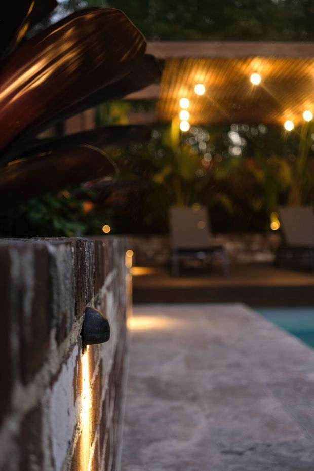 Moody close-up of a dark, textured masonry or stone pool wall with integrated warm linear lighting. A paved poolside terrace extends beside visible turquoise pool water, with lush planting and softly glowing outdoor lights in the blurred background. A large dark sculptural feature occupies the upper left.