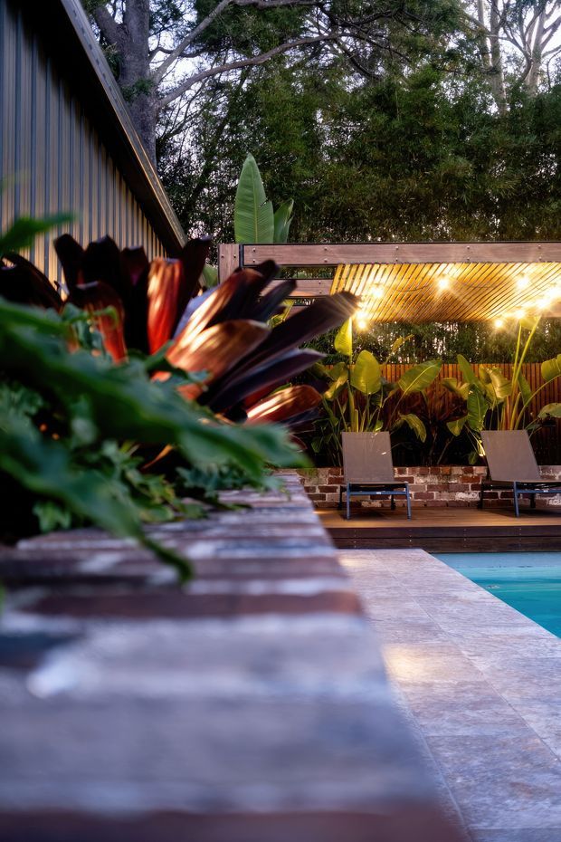 A lush landscaped poolside courtyard at dusk, with a timber-clad building along the left, a covered timber outdoor lounge or pergola lit with warm downlights, tropical potted planting, lounge chairs, brick detailing, and a turquoise swimming pool edged with stone paving in the foreground.