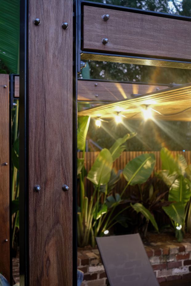 Close-up view of a dark timber-framed outdoor structure or cabinet with black metal edging, round metal fasteners, and reflective glazing. Warm integrated lighting illuminates a lush tropical courtyard garden with raised brick planters visible beyond. The image emphasizes the timber joinery, reflections, and atmospheric outdoor setting.