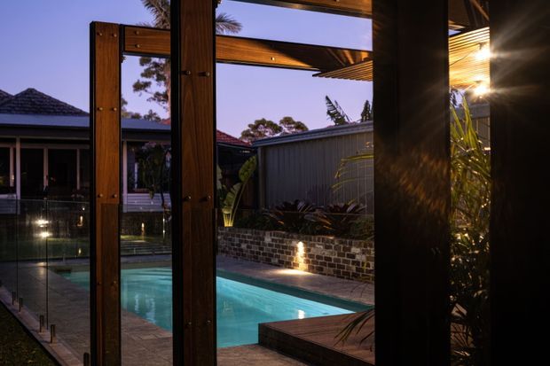 A dusk view from a sheltered outdoor living area through large timber-framed glazed doors toward a lit swimming pool. The scene includes a timber deck, brick planter wall, landscaped planting, corrugated metal fencing, and neighboring low-pitched tiled roof forms, with warm architectural lighting creating a relaxed atmosphere.