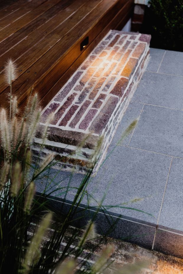 Close-up exterior detail of a landscaped patio or terrace, featuring a low curved bench or retaining wall faced with irregular reddish and cream brickwork. A dark timber-clad built-in structure with a small black recessed handle sits behind it, while large grey paving slabs and ornamental grasses complete the scene.
