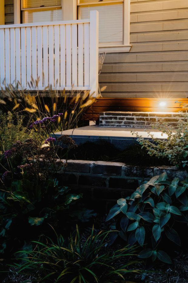 Close-up exterior view of a residential front entry, showing a white-painted porch balustrade, warm grey horizontal weatherboard cladding, windows with white trim, timber decking and steps, and layered garden planting bordered by dark masonry retaining walls. The scene is photographed in warm, low evening light.
