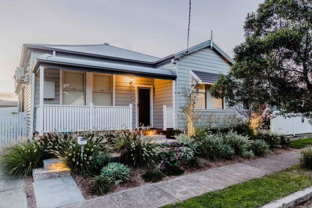 Street-facing exterior view of a small single-storey weatherboard house with a gabled front section, broad corrugated metal roof, enclosed front veranda, white picket-style balustrade and fence, and a landscaped garden with a concrete footpath. The facade is painted pale grey with white trims, and warm light glows from the entry and windows.