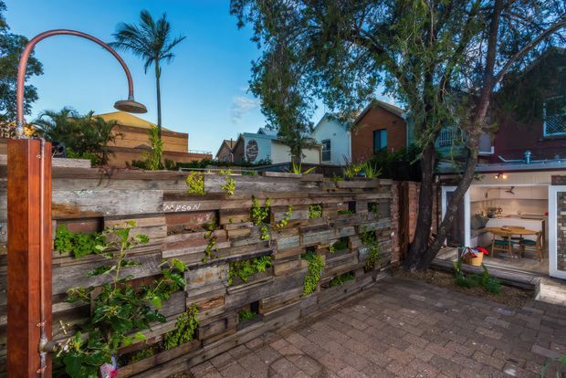 A residential outdoor courtyard or patio with weathered timber retaining walls and raised garden beds, climbing greenery, brick paving, and a distinctive curved copper-toned outdoor shower or tap fixture. Mature trees and palms surround the space, with neighbouring residential buildings visible beyond. A glazed opening on the right reveals an adjoining interior dining or living area.
