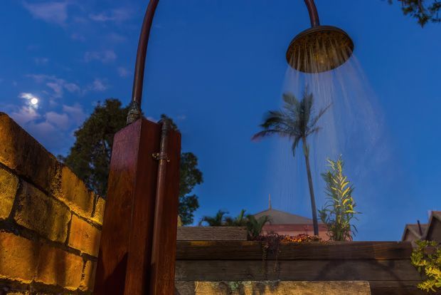 A dramatic dusk view of an outdoor shower with a large curved copper-toned pipe and round rainfall showerhead, set beside a rustic timber privacy fence and textured stone wall. Tropical palms, a tiled roof, and a deep blue evening sky form the background, with warm lighting highlighting the masonry and timber.