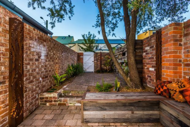 A compact enclosed brick courtyard or outdoor entertaining area with red brick boundary walls, paved flooring, timber screening and built-in timber bench seating. Mature trees and planting soften the space, while patterned outdoor cushions add colour. A small gate and adjoining rooftops are visible in the background.