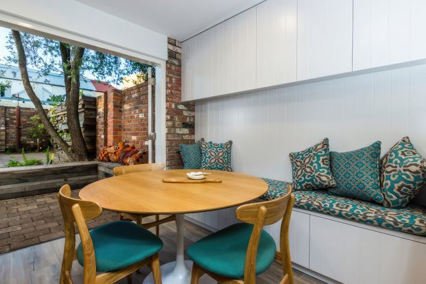 A compact, bright dining nook with a round timber dining table and matching timber chairs upholstered in teal. A built-in bench seat with patterned teal cushions sits beneath extensive white vertical-panel cabinetry. Exposed red brick walls and large glazing open the space toward a leafy courtyard, creating a blend of contemporary joinery and warm, textural materials.