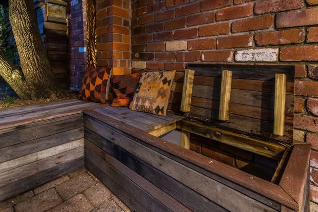 A rustic outdoor seating nook built against a red brick wall, featuring dark weathered timber bench seating, raised planter-style construction, timber backrest slats, and patterned woven cushions. A mature tree and surrounding paving reinforce the informal garden setting.