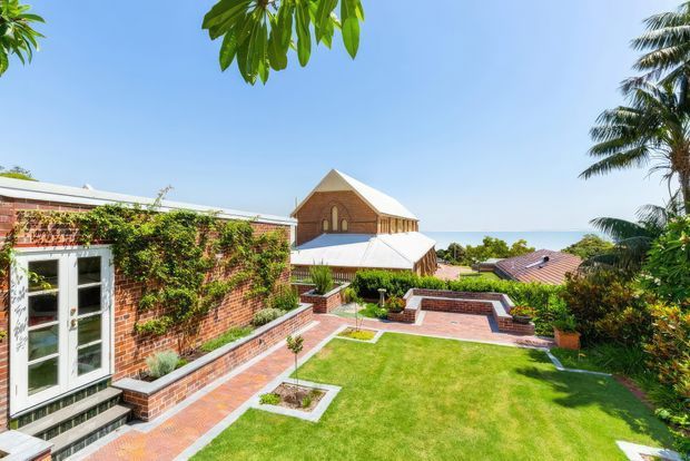 A bright landscaped courtyard and garden viewed from an elevated position, featuring red brick walls and paths, clipped lawn, planted borders, a white-framed glazed door, and a distinctive timber-clad gabled building with a light metal roof in the background. Tropical palms and blue sky surround the property, with a glimpse of the ocean beyond.