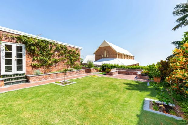 Bright landscaped courtyard garden beside a low red-brick building with white-framed glazed doors, climbing greenery, raised brick planters and paved paths. A distinctive gabled brick building with a white roof is visible beyond the courtyard, surrounded by manicured lawn and tropical planting.