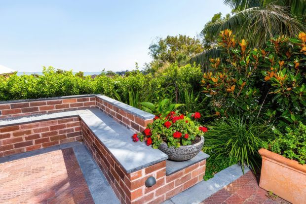 Sunny landscaped brick terrace or courtyard with built-in brick seating or planter walls capped in grey stone, a patterned brick-paved floor, and abundant tropical planting in containers and garden beds. A distant coastal or urban view is visible beyond the greenery.