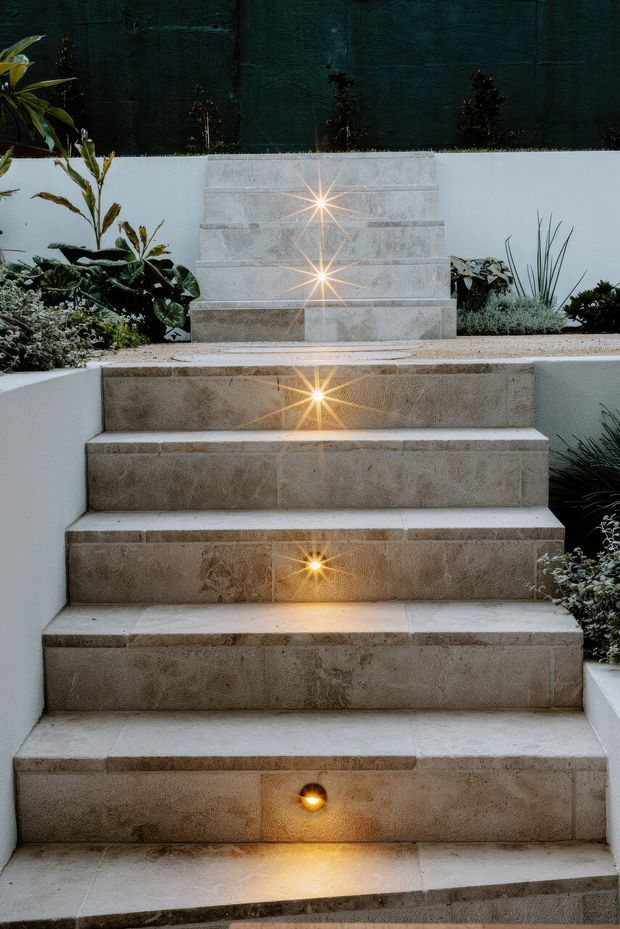 A landscaped exterior staircase rises through planted garden beds, finished in large warm beige stone tiles. Small integrated amber step lights are set into the stair risers, creating a strong illuminated pathway against white rendered retaining walls and dark screening above.