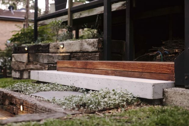 Close-up of a landscaped garden entry with layered concrete and weathered timber steps, built-in timber seating or edging, dark structural posts and railings, planted retaining walls, and small recessed step lights. The image emphasizes natural materials and a carefully integrated outdoor transition.