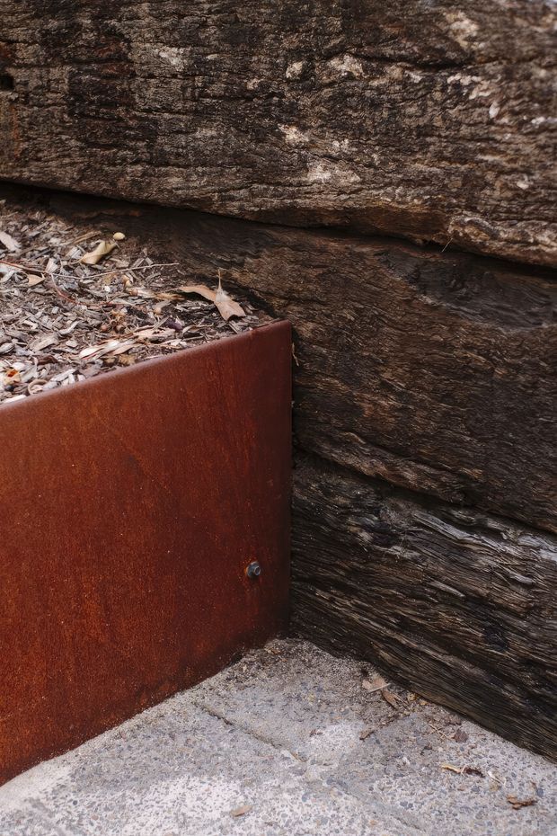 Close-up exterior detail of a rugged layered natural timber or log wall meeting a rusted weathering-steel planter or edging panel. The planter contains dry leaf litter, and the elements sit on a rough grey concrete or stone-paved surface. The image emphasises tactile weathered materials and a dark earthy palette.