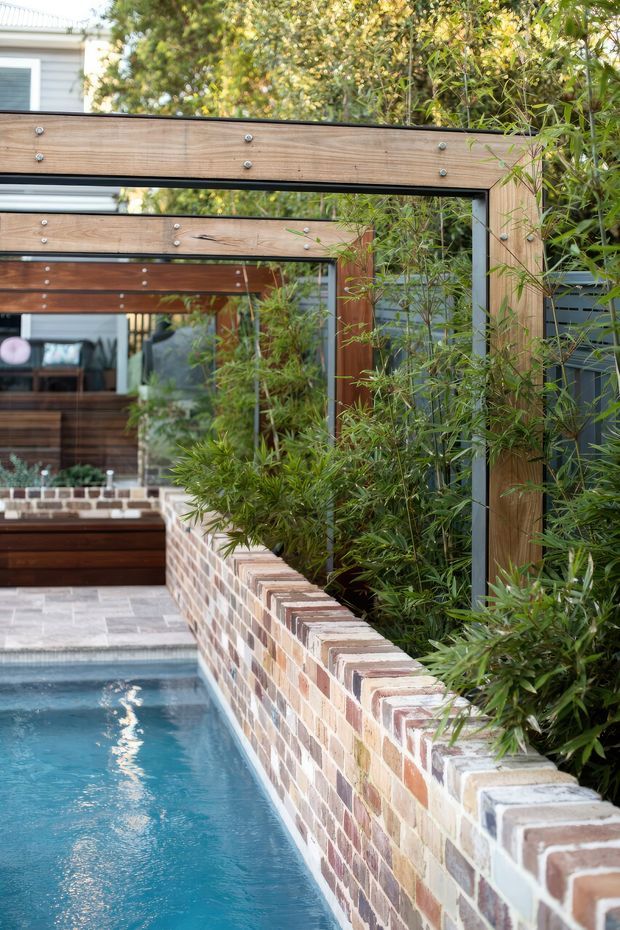 A narrow residential pool courtyard with blue water alongside a raised multi-coloured brick retaining wall and dense bamboo planting. Timber-framed horizontal screening and black metal supports create a layered architectural boundary, with a covered outdoor entertaining area and house visible beyond.