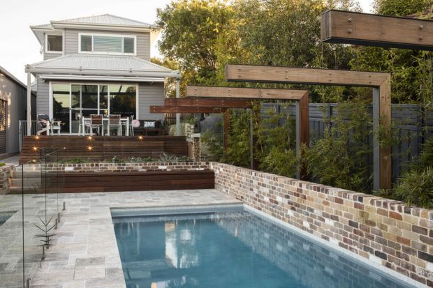 A landscaped backyard with a compact rectangular swimming pool, pale stone paving, a low exposed-brick retaining wall, and timber-framed garden screening. A raised timber deck with outdoor dining overlooks the pool and connects to a two-storey weatherboard house.