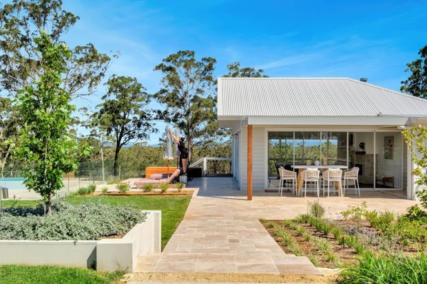 A bright single-storey contemporary home exterior with a pale corrugated pitched roof, light rendered walls, timber-toned posts, and extensive glazed doors opening onto a paved outdoor dining terrace. Landscaped raised garden beds and a lawn surround the house, with a fenced recreational area visible to the side.