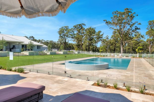 A bright outdoor pool area viewed from a shaded patio beneath a thatched umbrella. The scene features a large tiled swimming pool with a raised circular spa, frameless glass safety fencing, pale stone paving, landscaped garden edges, and a white single-storey house with a pitched metal roof in the background. Mature trees and open lawn surround the property.