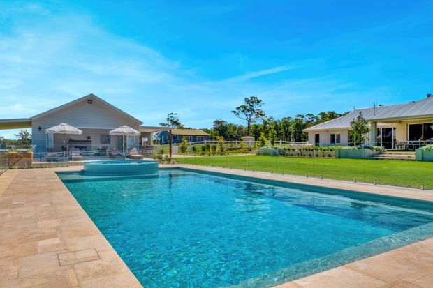 A bright outdoor view of a large rectangular swimming pool with turquoise water, surrounded by tan tiled paving. A small poolside pavilion with umbrellas sits at the far end, with manicured lawn, low fencing, and single-storey light-coloured buildings with pitched metal roofs in the background.