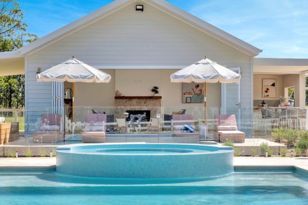 A bright exterior view of a white weatherboard-style pool house or pavilion opening onto a covered outdoor living area. The foreground features a turquoise swimming pool with a raised circular spa, while the pavilion includes glass balustrading, striped umbrellas, outdoor seating, and a visible bar or lounge area.