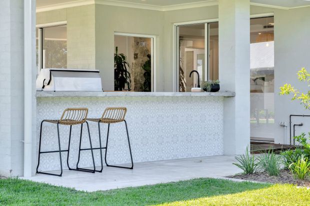 Covered outdoor entertaining area with a built-in bar counter and patterned tile-clad front, two bar stools, a barbecue, and a sink. White rendered columns and walls frame glazed openings into the house, with landscaped planting and lawn in the foreground.