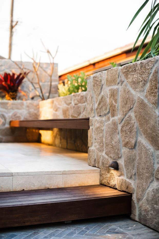 A warm, sunlit outdoor courtyard or terrace with stepped levels, featuring rough natural stone retaining walls, dark timber steps and edging, and pale tiled paving. A low timber bench or platform extends along the stone wall, with tropical and ornamental planting around the perimeter. Small integrated wall lighting adds a subtle accent.