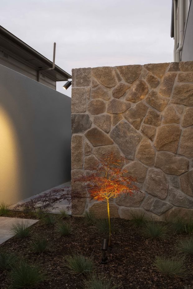A narrow landscaped courtyard or side passage beside a building, featuring a tall irregular natural-stone masonry wall in warm brown and taupe tones. A smooth dark grey rendered wall and roof edge sit to the left, with a small spotlight illuminating an ornamental tree and low grasses in a mulched garden bed. The scene has a refined, subdued contemporary exterior character under an overcast sky.
