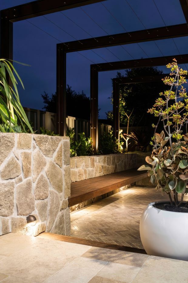 A landscaped outdoor courtyard or garden terrace at dusk, featuring dark steel-framed openings or pergola-like structures, illuminated planting, rustic stone retaining walls, a built-in timber bench, patterned paving, and a large white planter. The space has a warm, layered atmosphere with strong architectural framing and garden lighting.