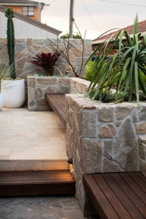 A landscaped residential courtyard or terrace with raised stone masonry planters, integrated dark timber decking and bench seating, pale stone-paved flooring, and tropical plants. The view includes neighbouring residential rooflines and a timber privacy screen, with warm integrated lighting along the deck and planter edges.
