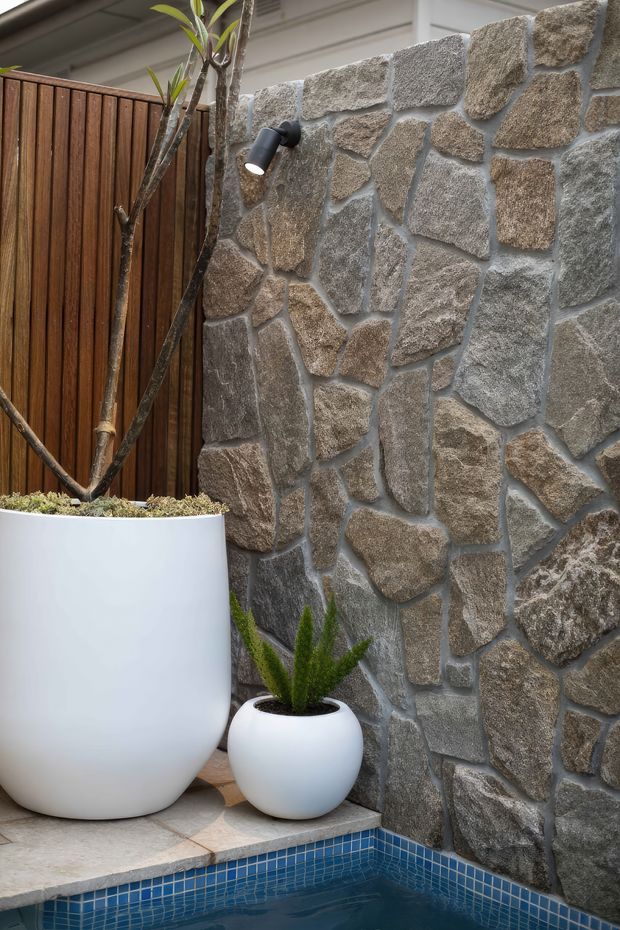 A close-up view of a refined outdoor courtyard pool corner. A rugged irregular-stone feature wall is paired with warm vertical timber screening, black wall-mounted lighting, white sculptural planters, and a blue tiled pool edge.