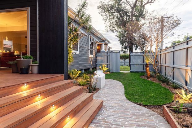 A landscaped residential side courtyard and entry path beside a dark weatherboard-clad home. Broad timber steps with integrated warm lighting lead to an open glazed doorway, while a curved brick-paved walkway passes through lawn and planted garden beds toward a timber-and-metal boundary fence. The scene has a warm, welcoming contemporary character with subtle coastal or Australian suburban cues.