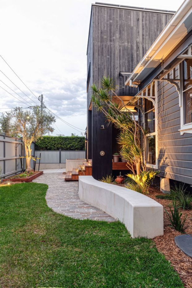 A landscaped side courtyard and entry path beside a residential house. The home combines dark vertical timber cladding on a tall facade with grey weatherboard siding, white-trimmed windows, and a covered timber-framed porch. A curved rendered concrete retaining wall borders a paved path, with ornamental grasses, yucca-like planting, lawn, raised garden beds, and a gated side boundary.