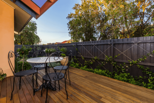 A warm outdoor timber deck adjoining a house, furnished with a small wrought-iron dining table and chairs. The deck is enclosed by a dark timber privacy fence with climbing greenery, while mature trees and a clear sky are visible beyond. The house has warm painted walls and exposed roof eaves.