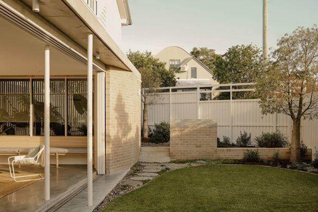 A contemporary residential courtyard viewed from a covered outdoor living area. The home has pale brick walls, slim white columns, timber-toned decking, and large glazed openings with vertical screening. A neatly landscaped lawn, stepping-stone path, raised brick planter and lightweight metal boundary fencing define the compact garden.
