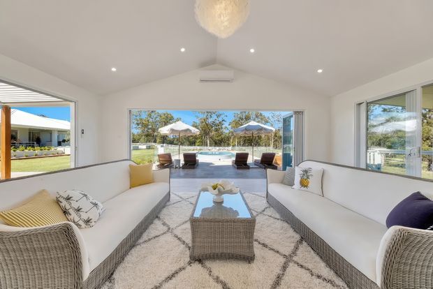 Bright, airy sunroom-style living space with white wicker sofas, a glass-topped wicker coffee table, and a neutral patterned rug. Large windows and wide sliding doors open toward a pool and landscaped outdoor area, while recessed ceiling lights and a sculptural pendant provide illumination. The room has a clean coastal-resort feel with strong indoor-outdoor connection.