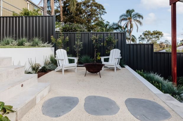 A landscaped outdoor terrace with a pale textured paved surface, large irregular stepping stones, white Adirondack-style chairs, and a small dark metal fire pit. Black vertical metal fencing encloses the space, with raised white rendered garden beds, ornamental grasses, climbing plants, and mature tropical and native trees beyond. Concrete steps connect to the terrace on the left.