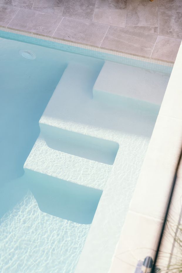 Close-up view of a pale blue swimming pool with broad submerged entry steps and integrated seating or ledges. The pool has a light tiled coping and surrounding pale stone or ceramic paving, with bright sunlight creating rippled reflections across the water.