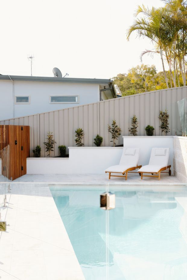 A bright outdoor swimming pool courtyard with pale tiled paving, white rendered walls and raised planters, a vertical-panel privacy fence, timber gate, and two white cushioned sun loungers. A low flat-roofed building and tall palms are visible behind the fence.