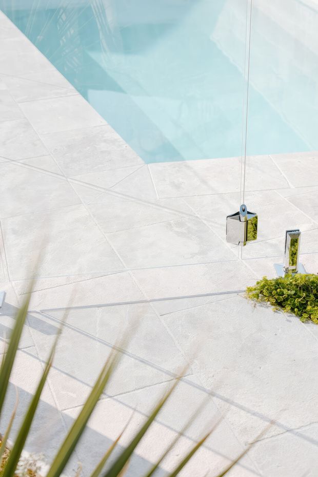 A bright outdoor pool area with pale large-format tiled paving surrounding clear turquoise water. Small chrome fixtures and low green planting sit along the pool edge, with strong sunlight and crisp shadows creating an airy contemporary atmosphere.