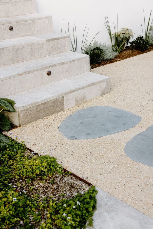 A landscaped outdoor garden pathway with broad pale stone steps, integrated round step lights, a light-coloured granular paving surface, and irregular dark grey stepping stones. Low groundcover planting and drought-tolerant ornamental plants border the path against a white wall.